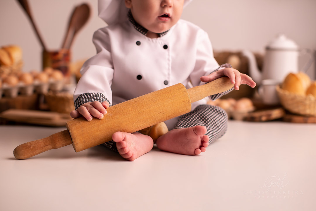 ensaio de bebe 9 meses padeiro com paes em cestos colheres de pau bule de cafe ovos e bebe com roupa de chef de cozinha  segurando rolo de massa de madeira