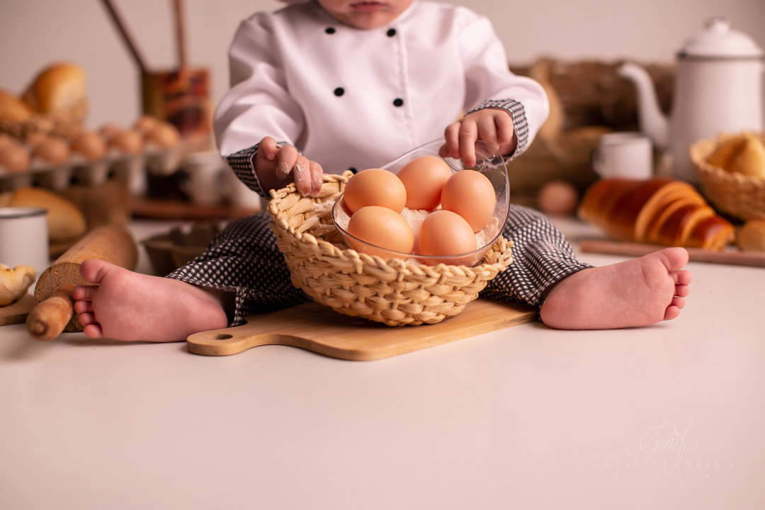 ensaio de bebe de 9 meses de padaria com roupa de chef de cozinha  segurando um cesto de ovos de galinha
