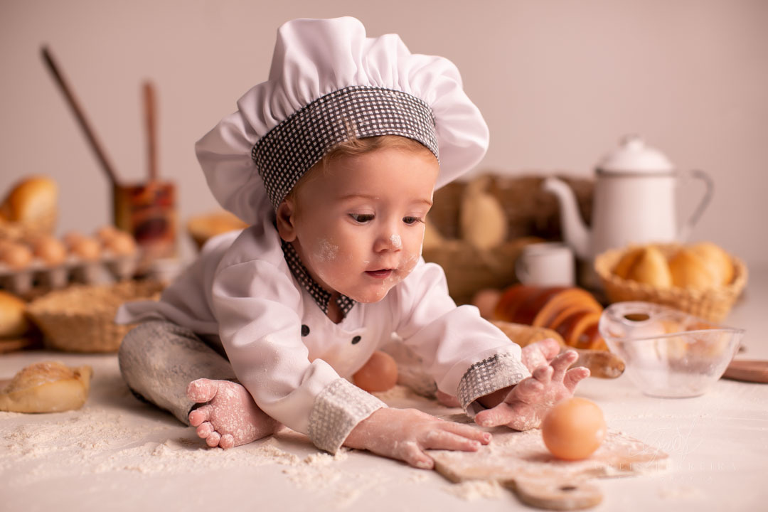 ensaio de bebe de 9 meses de padaria segurando uma tabua de madeira suja de farinha