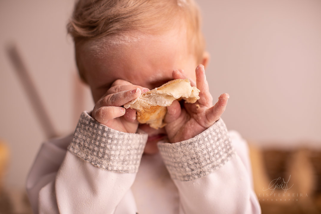 ensaio de bebe 9 meses padeiro com paes em cestos colheres de pau bule de cafe ovos e bebe com roupa de chef de cozinha segurando um pao em frente aos olhos