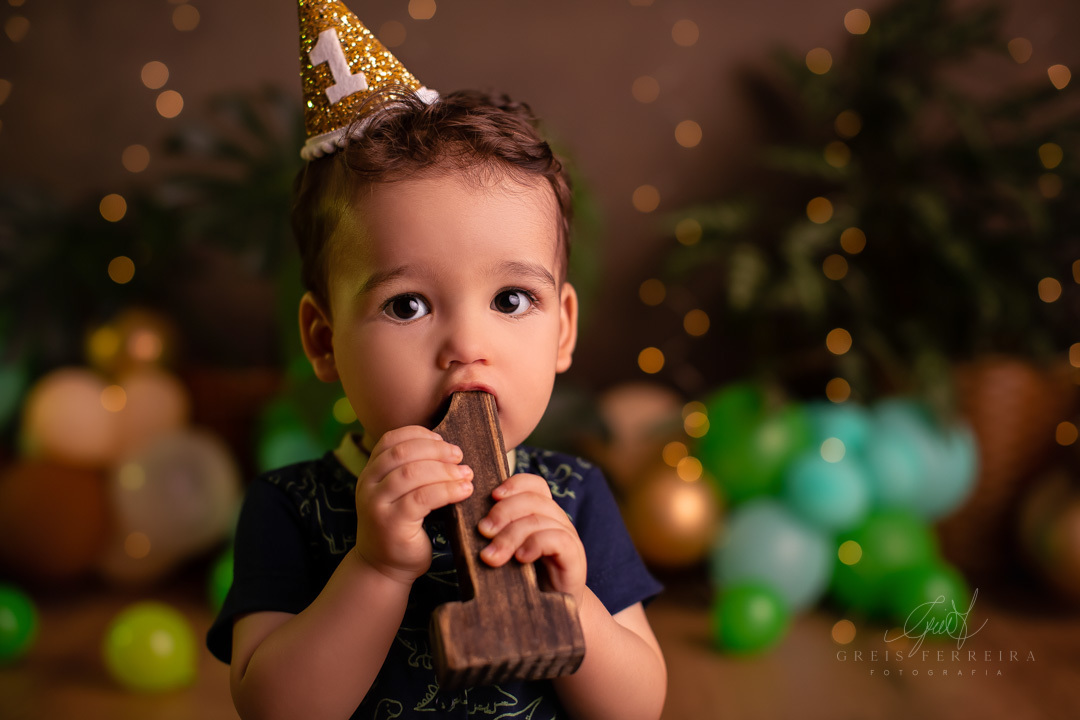 bebe com o numero 1 em madeira na boca  usando chapeu de festinha de aniversario ao fundo temos baloes plantas naturais e pisca-pisca