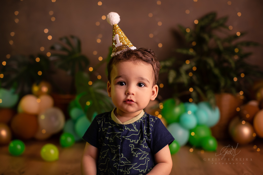 foto de meio plano de bebe usando chapeu de festinha de aniversario ao fundo temos baloes plantas naturais e pisca-pisca