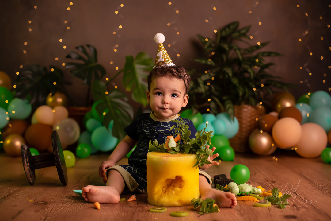 smash the fruit de dinossauro com um bolo de melancia amarela decorado com kiwi hortela manga dinossauros de biscuit em pedestal de madeira a frente de um bebe usando chapeu de festinha de aniversario com fundo temos baloes plantas naturais e pisca-pisca