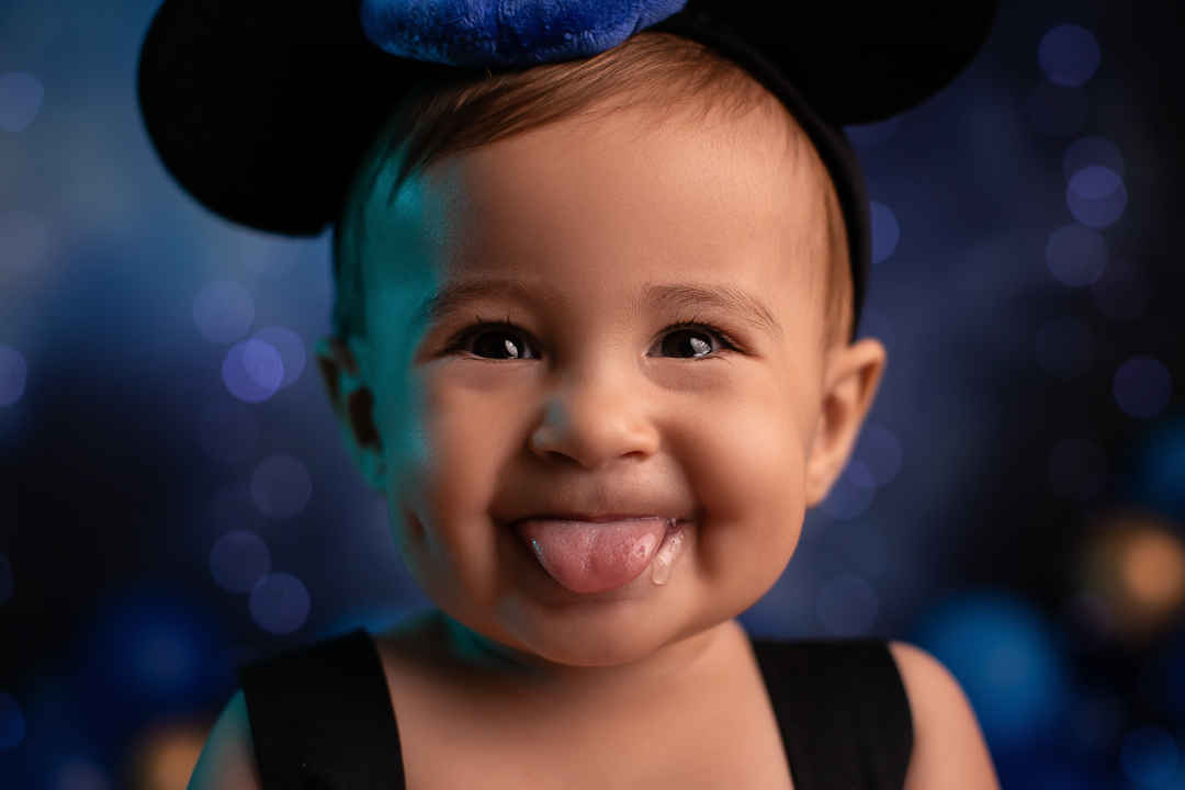 Smash the cake mickey ensaio de bebe chapeu de feiticeiro com orelhas do mickey na cabeça de um bebe colocando a lingua para fora com fundo azul piscas e baloes desfocado