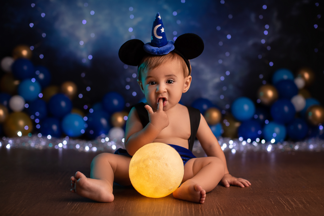 Smash the cake mickey ensaio de bebe chapeu de feiticeiro com orelhas do mickey na cabeça de um bebe com o dedo na boca sentado atras de uma lua acesa entre as pernas com fundo azul desfocado