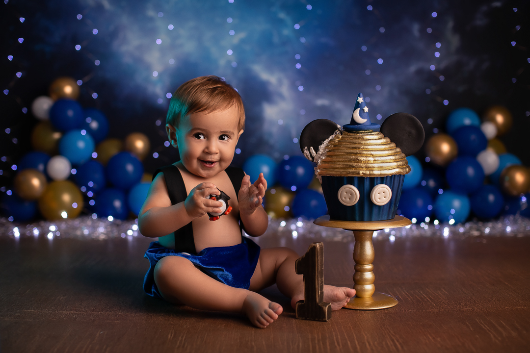 Smash the cake mickey ensaio de bebe com bolo de chapeu de feiticeiro com orelhas do mickeycom bebe sentado no chao sorrindo com  com fundo azul piscas e baloes desfocado