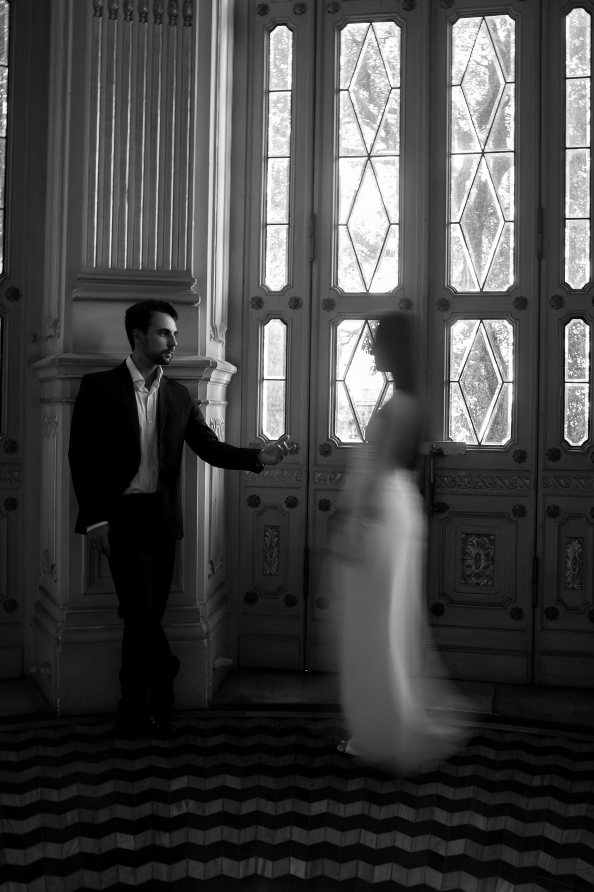 Fotografia artística a preto e branco. Noivo encostado a uma porta clássica de madeira e vidro, enquanto a noiva passa num movimento romântico e desfocado. Fotografia de casamento criativa.