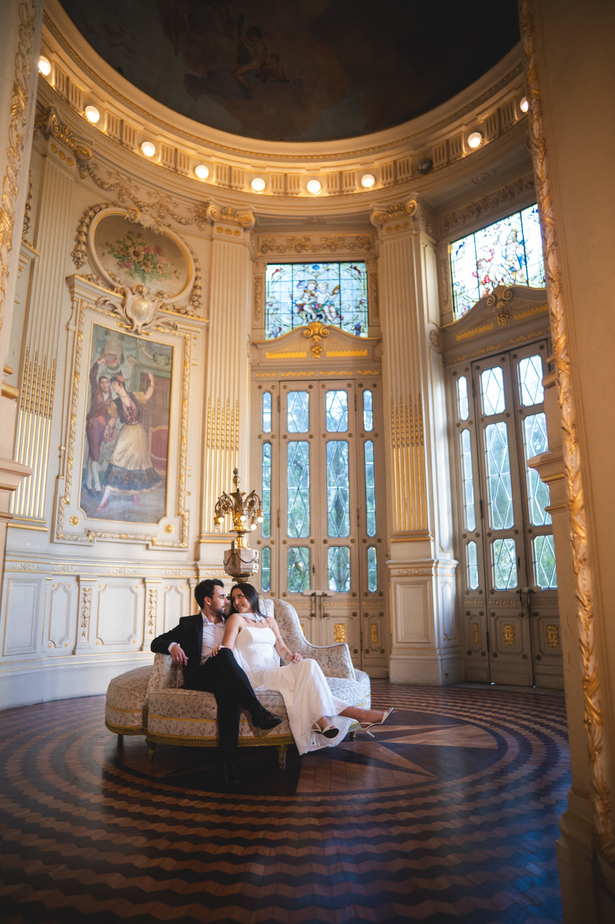 Casal de noivos sentados num sofá clássico no imponente Theatro Municipal do Rio de Janeiro. Fotografia de casamento com luz natural, vitrais e cenário de luxo.