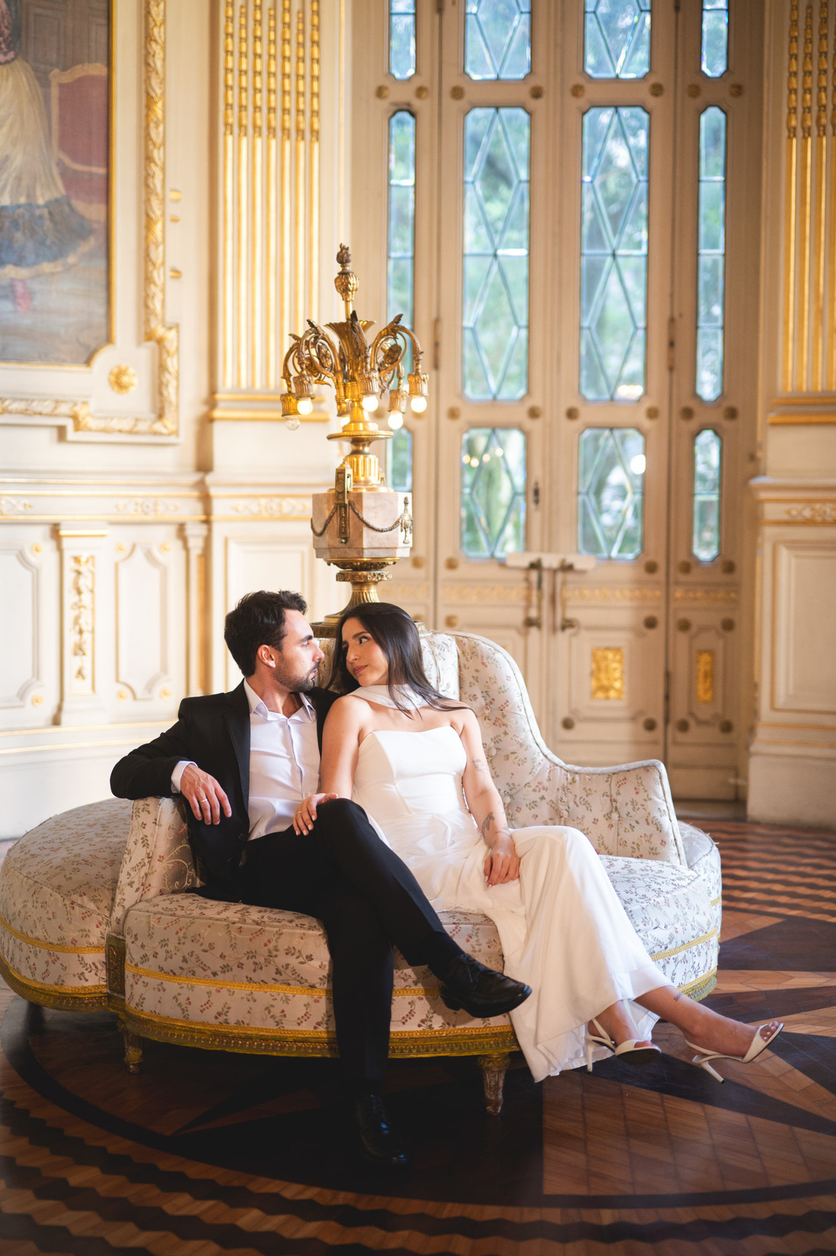 Noivos Maria Eduarda e Caio abraçados num luxuoso sofá de época. Sessão fotográfica pré-casamento no Rio de Janeiro com luz suave e tons quentes.
