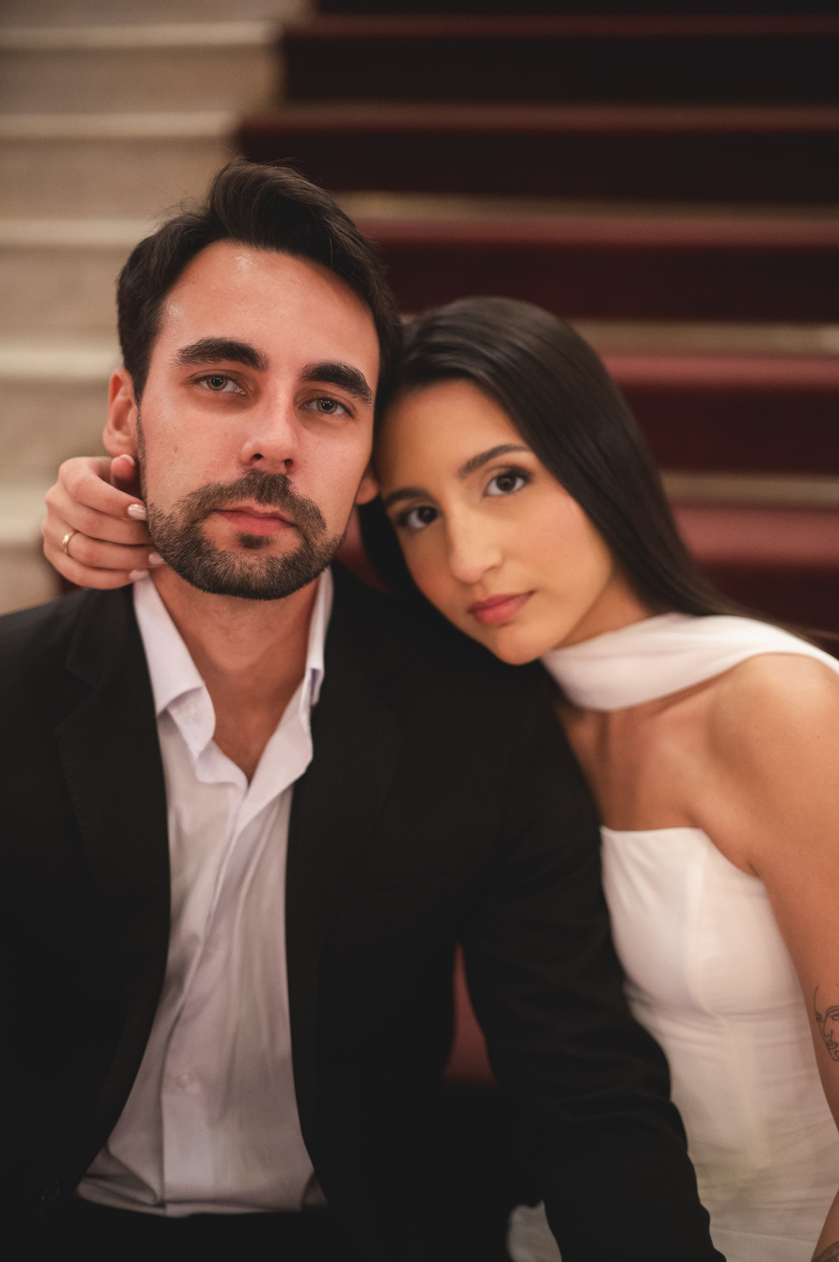 Retrato elegante em cores de uma noiva com um olhar direto, usando um vestido branco cai-cai, realçando sua tatuagem no braço e sua expressão refinada contra o fundo luxuoso e ornamentado do Theatro Municipal, Rio de Janeiro