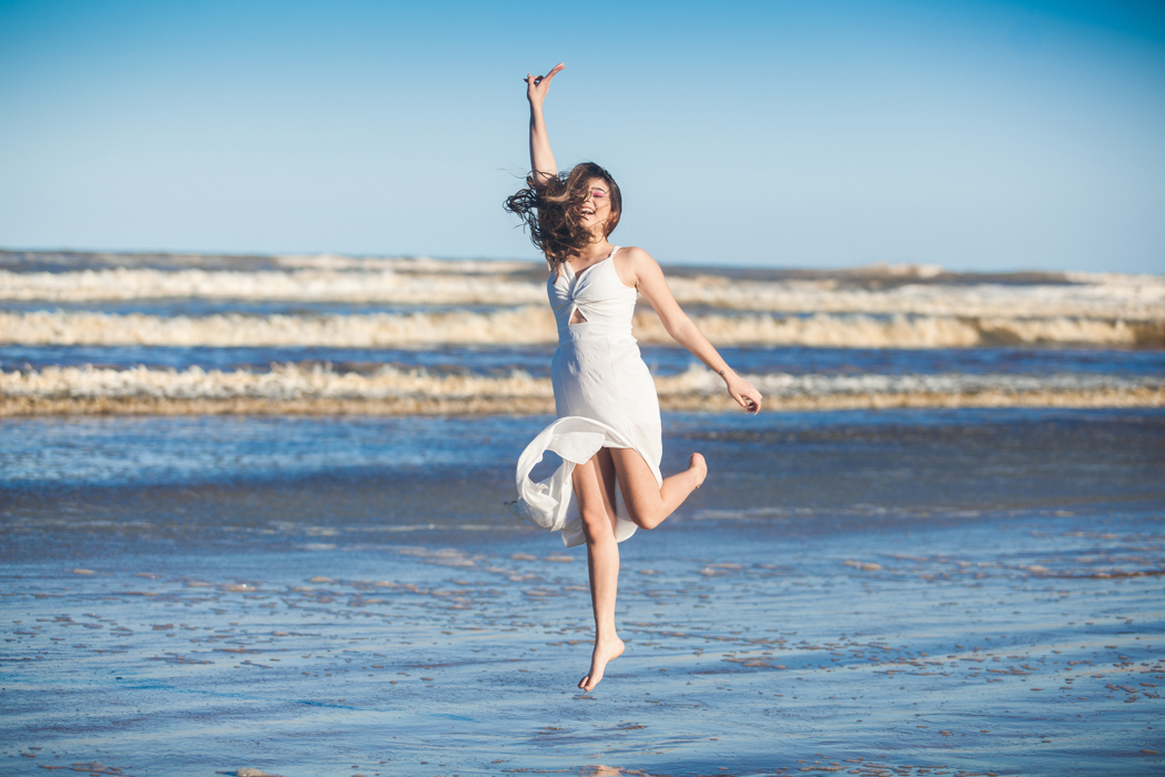 jovem de vestido branco saltando num pé só na beira do mar