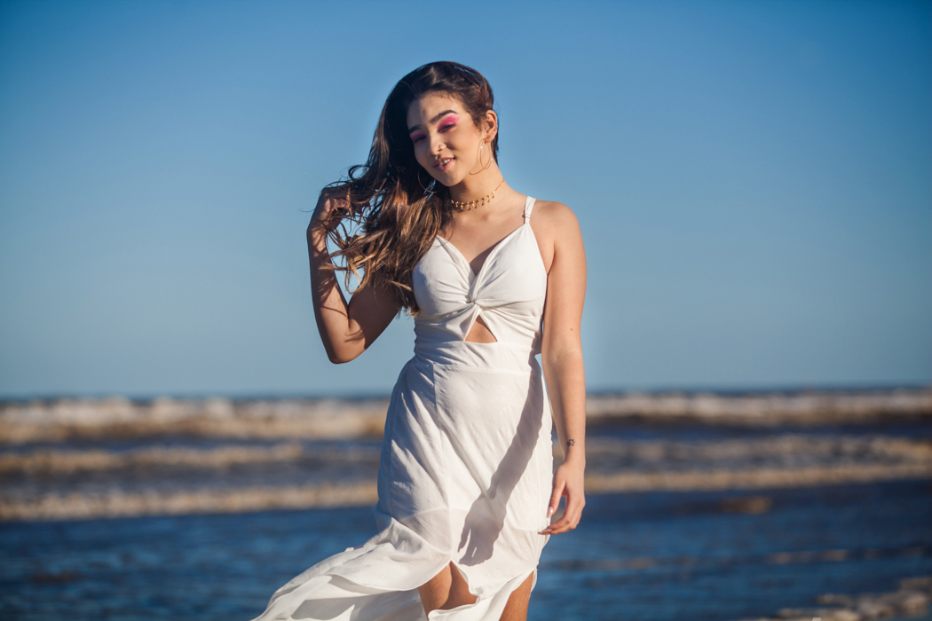 jovem com mão no cabelo e vestido branco balançando ao vento na bera da praia