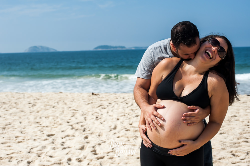 casal rindo e abraçados durante ensaio fotografico de gestante com a praia do leblon ao fundo