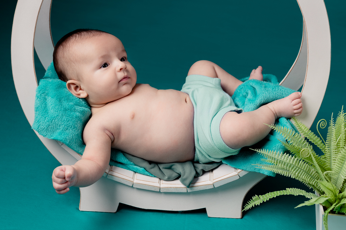 Foto de bebê menino deitado, em estúdio, com fundo verde, cama em arco e planta. Foto por Ander Lopes Fotografia, Taquara.