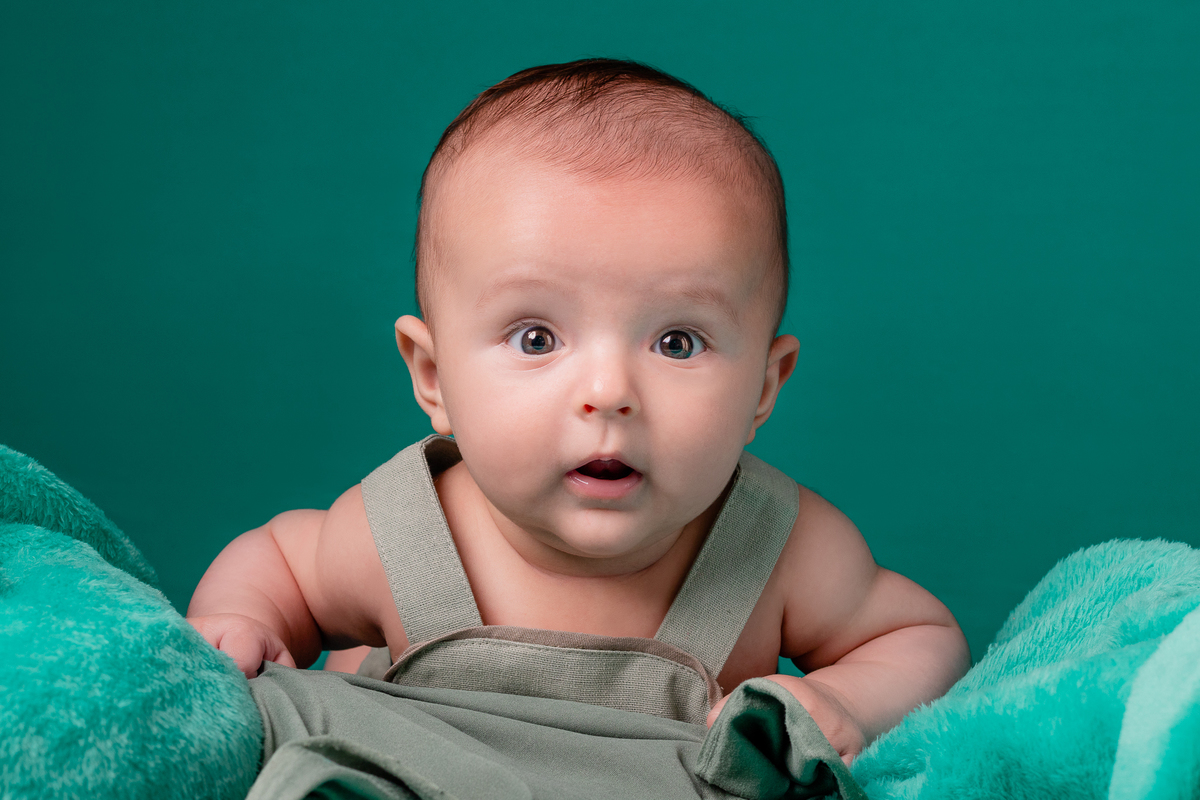 Foto de close de bebê menino, em estúdio, com fundo verde, cama em arco e olho brilhando. Foto por Ander Lopes Fotografia, Taquara.