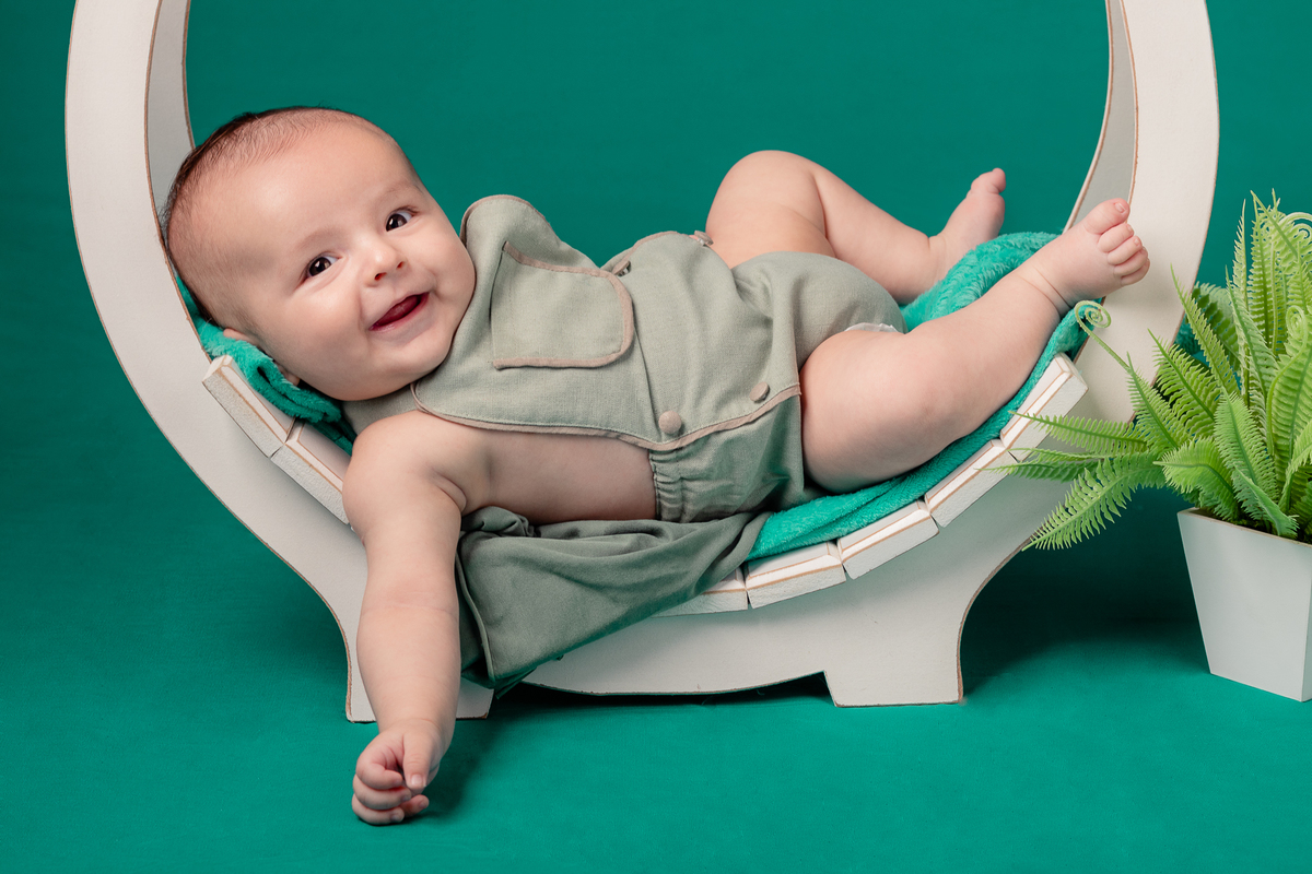 Foto de bebê menino sorrindo, em estúdio, com fundo verde, cama em arco e planta. Foto por Ander Lopes Fotografia, Taquara.