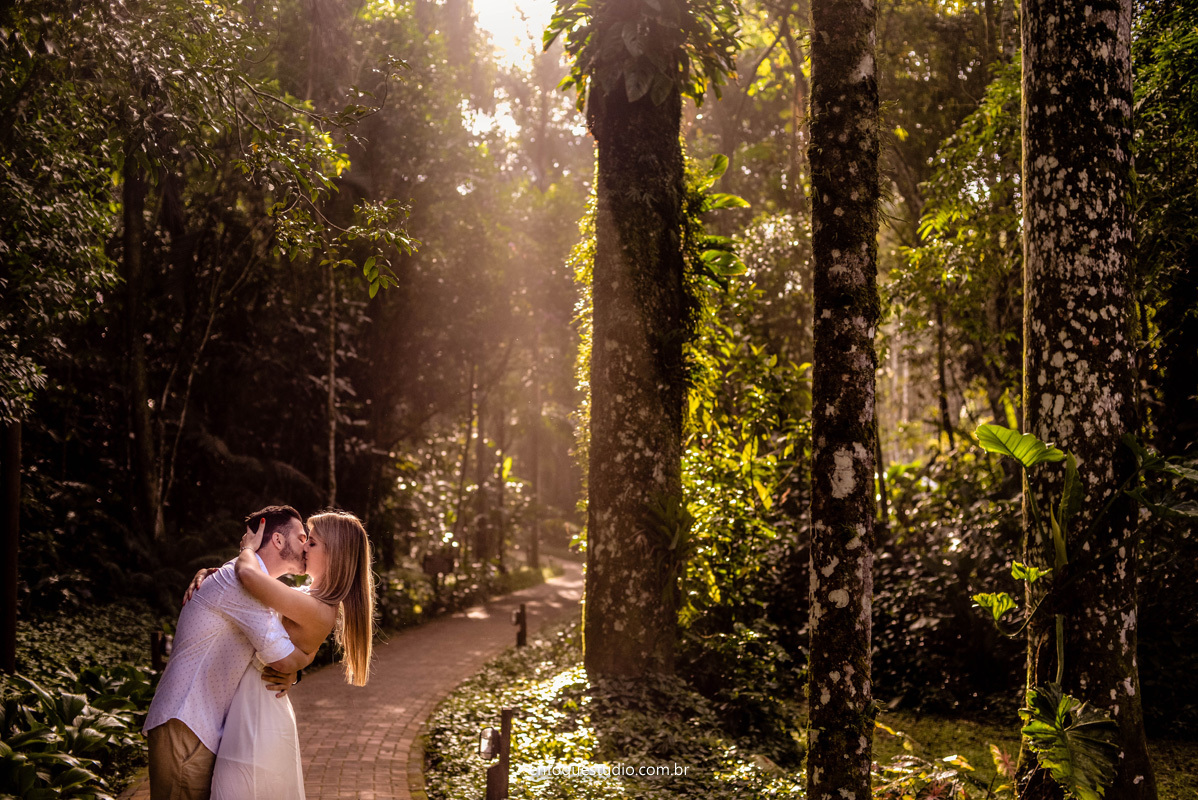 Foto Casal se beijando em um caminho com luz do sol entrando ao fundo e uma linda área verde
