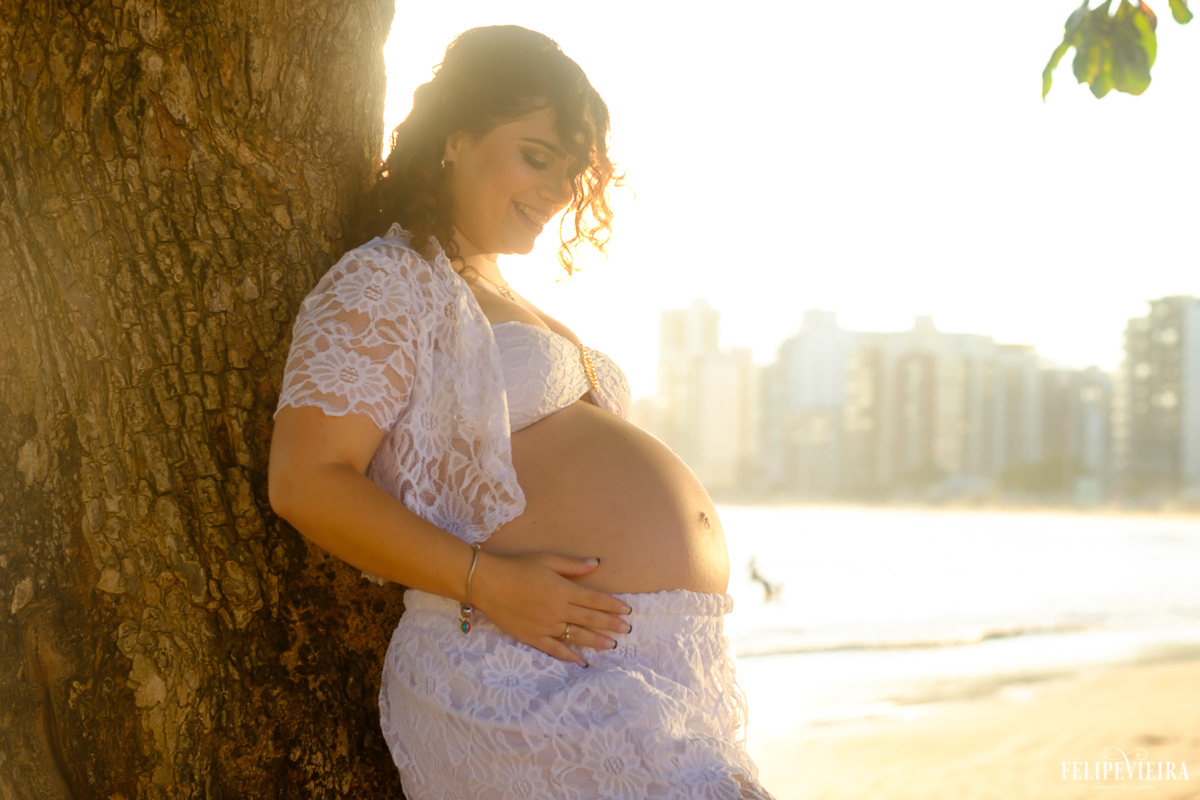gestante com roupa branca apoiada na árvore em frente à praia foto feita por felipe vieira fotógrafo de gestantes
