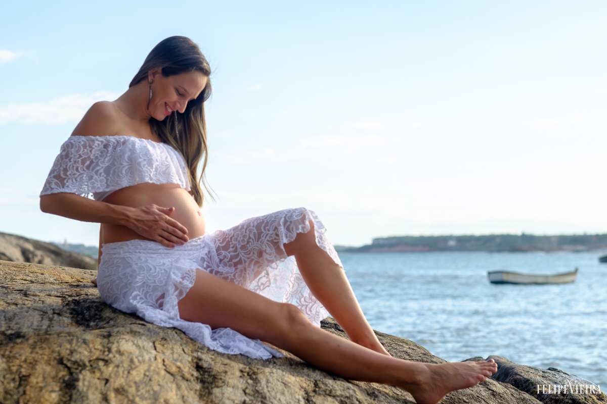 gestante sentada nas pedras com o mar em um belo céu azul ao fundo foto feita por felipe vieira fotógrafo de gestantes
