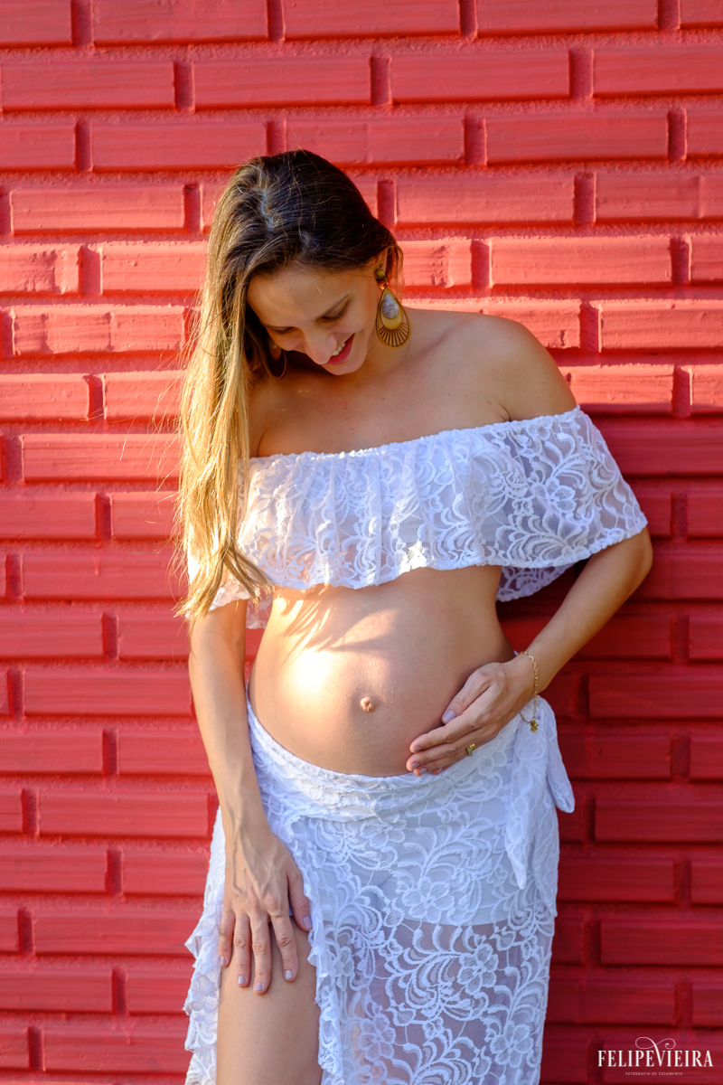 gestante em frente a um muro vermelho vestida de branco e olhando para a barriga feliz foto feita por felipe vieira fotógrafo de gestantes