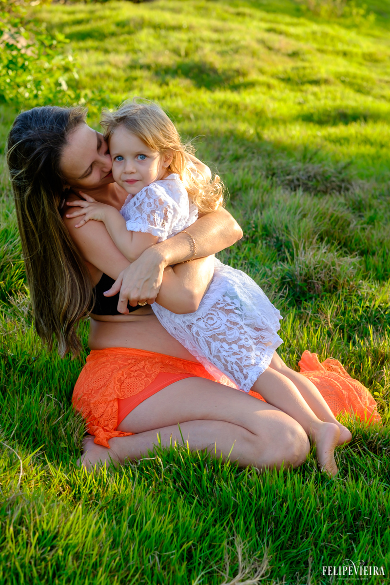 mãe abraçando sua filha sentada na grama feliz pelo próximo filho por vir foto feita por felipe vieira fotógrafo de gestantes