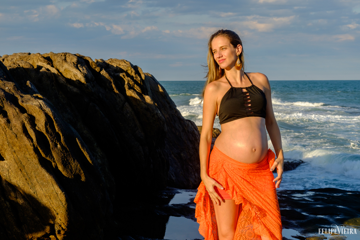 gestante em frente às pedras e ao mar com um belo pôr do sol começando a aparecer foto feita por felipe vieira fotógrafo de gestantes