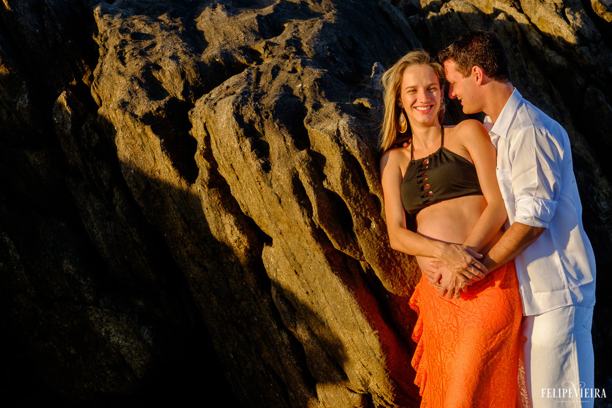 marido e esposa gestante abraçados e sorrindo em frente às pedras na praia foto feita por felipe vieira fotógrafo de gestantes