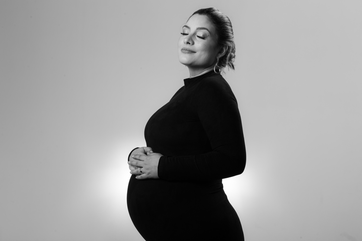 Book de gestante em estúdio com fundo escuro e luz suave. Sessão de fotos de grávida com postura relaxada e confiante.