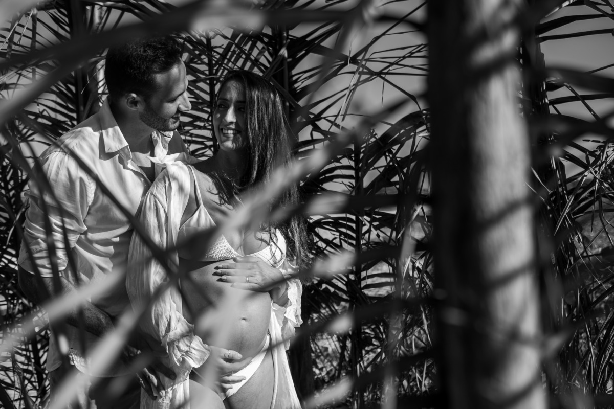 Fotografia de gestante com elementos da natureza à beira-mar. Gestante com o marido em meio a folhagens de coqueiros, fotografia em preto e branco