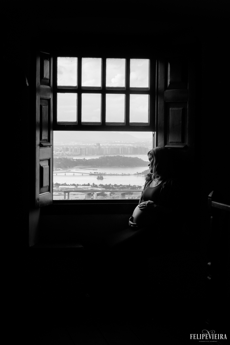 foto em preto e branco de gestante na beira da janela com vitória ao fundo foto feita por felipe vieira fotógrafo de gestantes