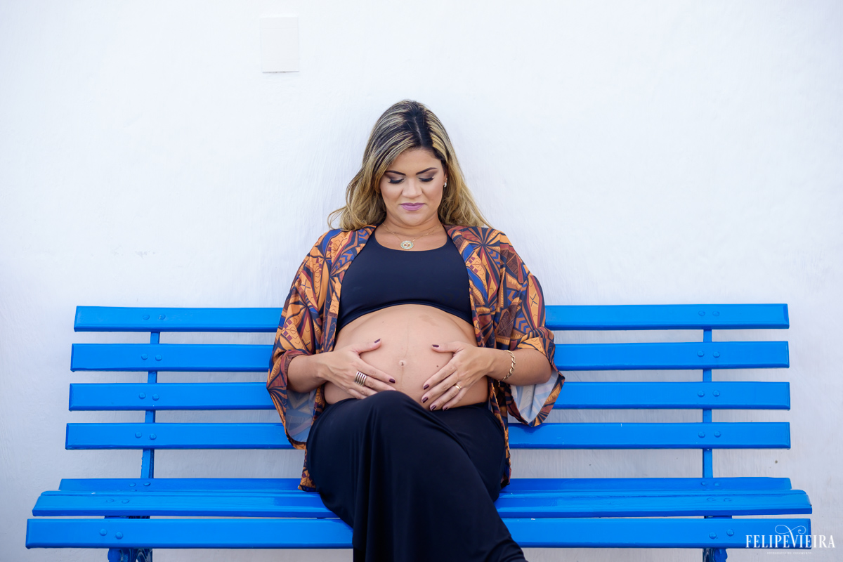 gestante sentada em um banco azul com as mãos na barriga e a olhando com felicidade foto feita por felipe vieira fotógrafo de gestantes