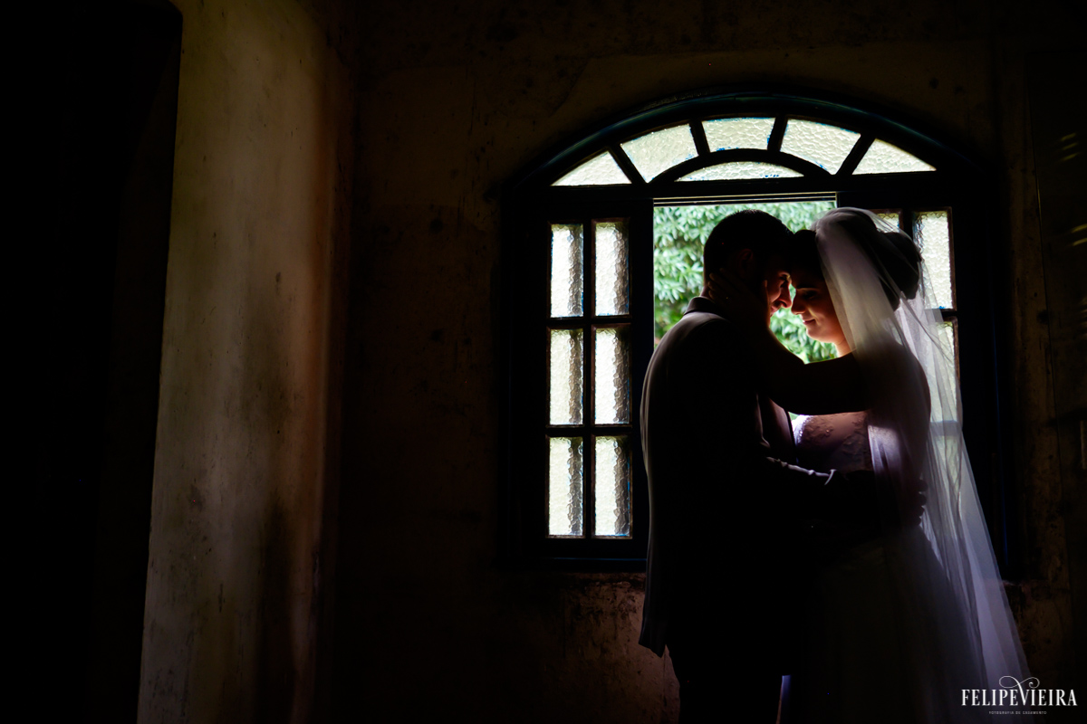 Noivos em tons escuros em frente a uma janela em foto externa foto feita por felipe vieira fotografo de casamentos em guarapari
