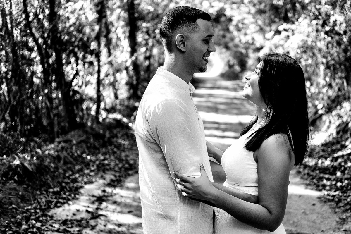 Casal sorrindo de frente um para o outro durante ensaio pré-casamento em Guarapari ele veste bata branca ela vestido branco em um caminho arborizado foto preto e branco destacando o momento romântico natural foto por fotógrafo especializado em casamentos