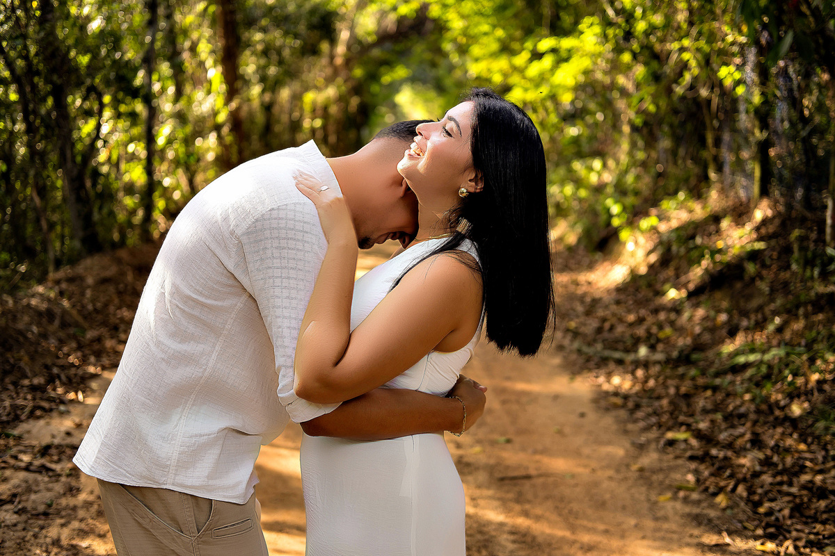 Casal abraçado em caminho arborizado durante ensaio pré-casamento em Guarapari ele veste bata branca ela vestido branco ele encosta a cabeça no ombro dela foto ao ar livre por fotógrafo especializado em casamentos destacando o amor