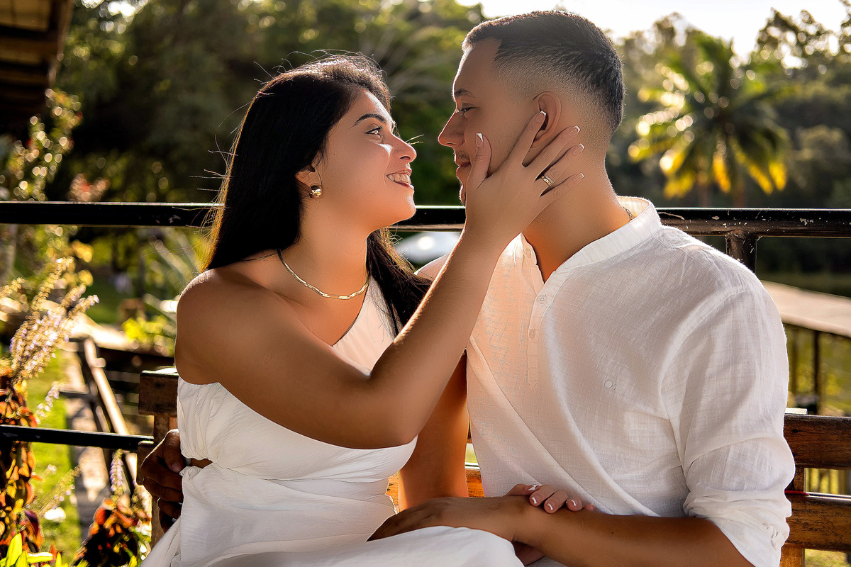 Casal sentado de mãos dadas durante ensaio pré-casamento em Guarapari ele veste bata branca ela vestido branco eles se olham nos olhos foto ao ar livre por fotógrafo especializado em casamentos