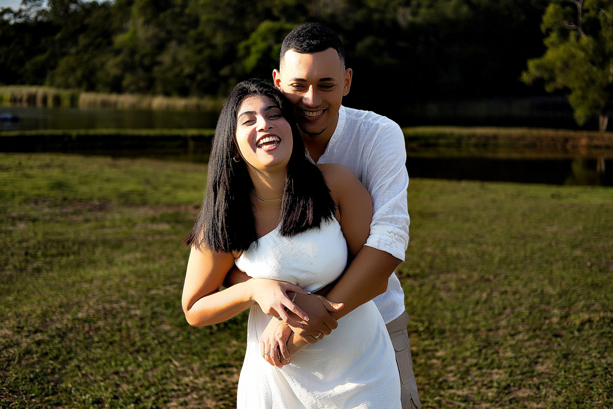 Casal rindo juntos em área gramada durante ensaio pré-casamento em Guarapari ele veste bata branca ela vestido branco ele a abraça por trás foto ao ar livre por fotógrafo especializado em casamentos