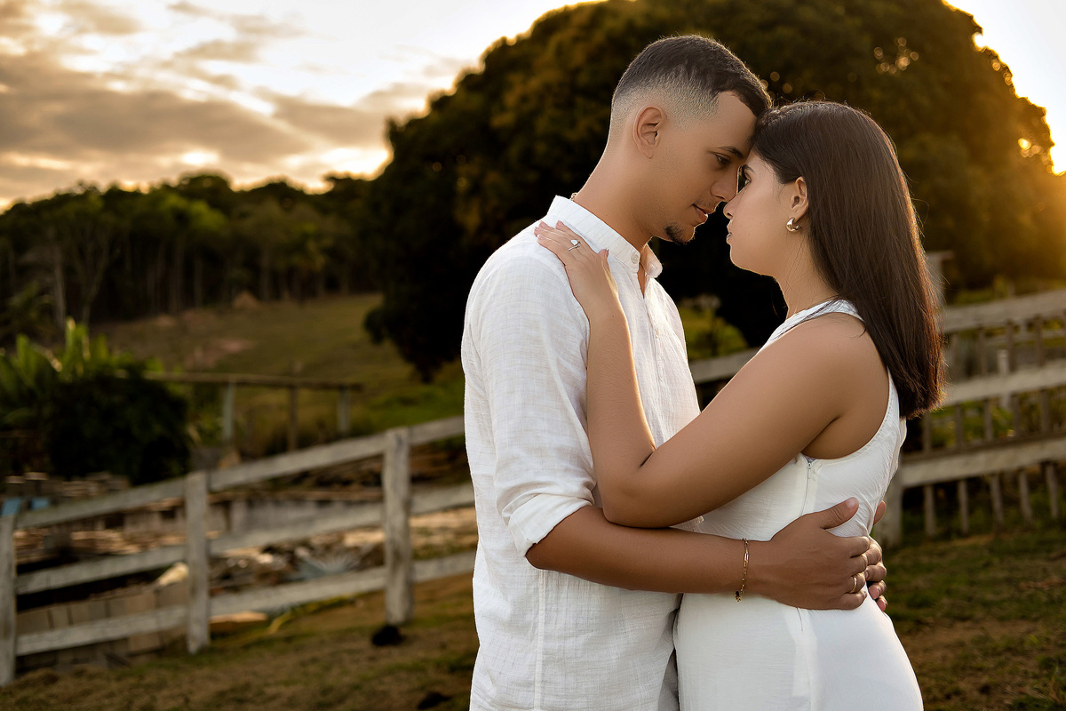 Casal abraçado ao pôr do sol durante ensaio pré-casamento em Guarapari ele veste bata branca ela vestido branco eles se olham com ternura foto ao ar livre por fotógrafo especializado em casamentos