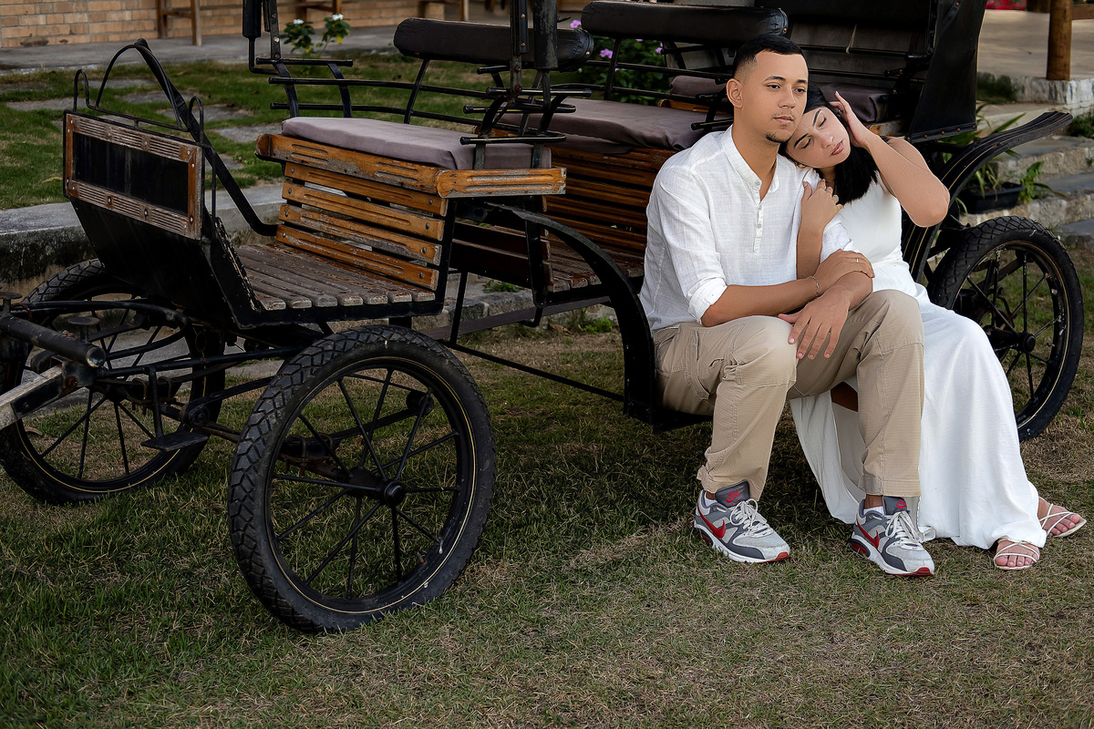 Casal em ensaio pré-casamento em Guarapari sentado em frente a uma carruagem antiga Ele veste camisa branca e calça bege ela usa vestido branco Ela encosta a cabeça no ombro dele mostrando carinho Foto ao ar livre por fotógrafo especialista em casamento