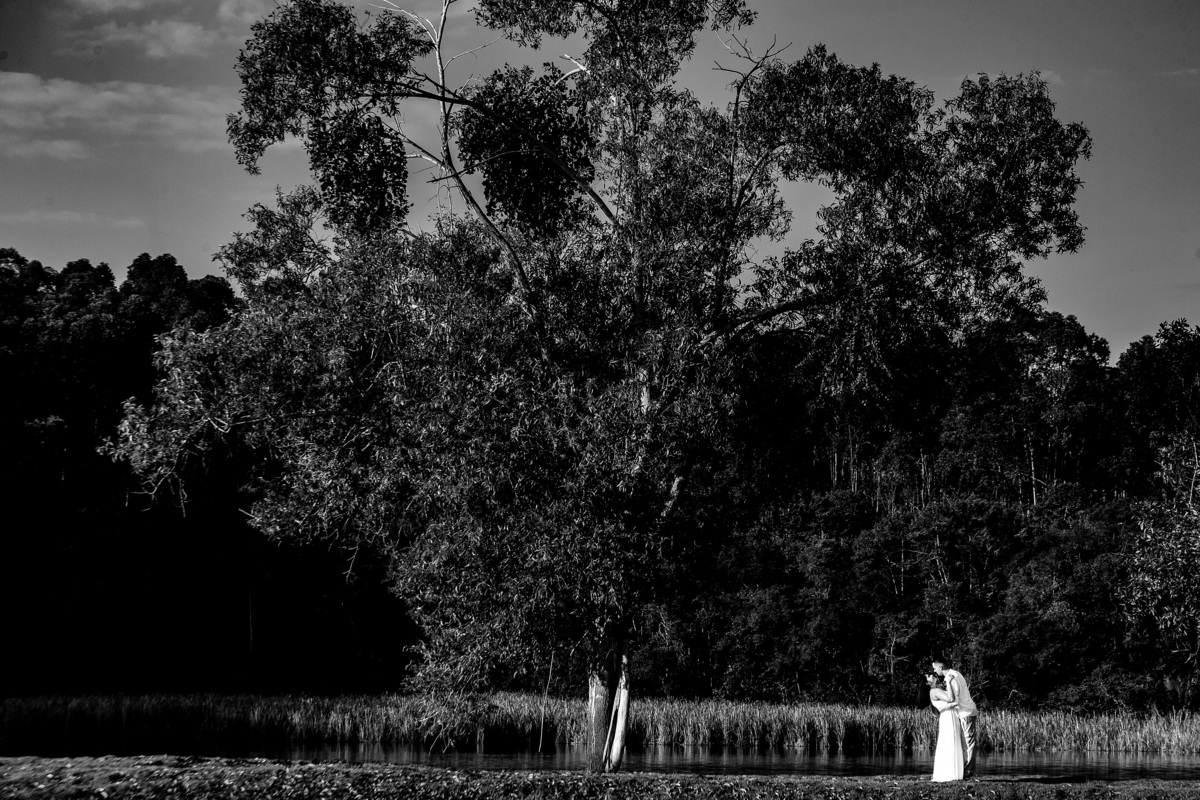 Casal abraçado à beira de um lago cercado por árvores em ensaio pré-casamento em Guarapari ele veste bata branca ela vestido branco foto preto e branco destacando a beleza natural e o romance por fotógrafo especializado em casamentos