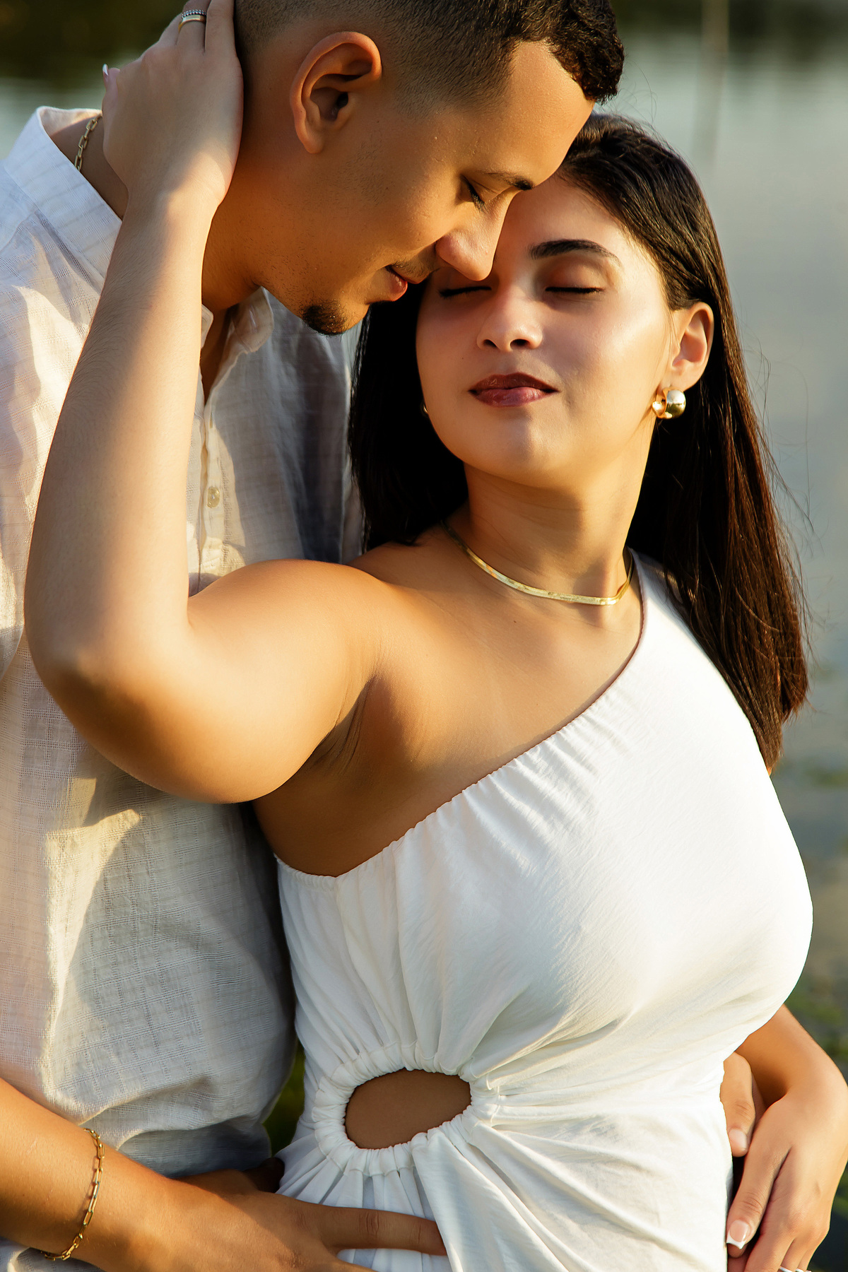 Casal em abraço próximo à beira do lago com vegetação alta ao fundo em ensaio pré-casamento em Guarapari; ele veste bata branca, ela vestido branco; luz suave do final de tarde; fotógrafo especializado em casamentos.