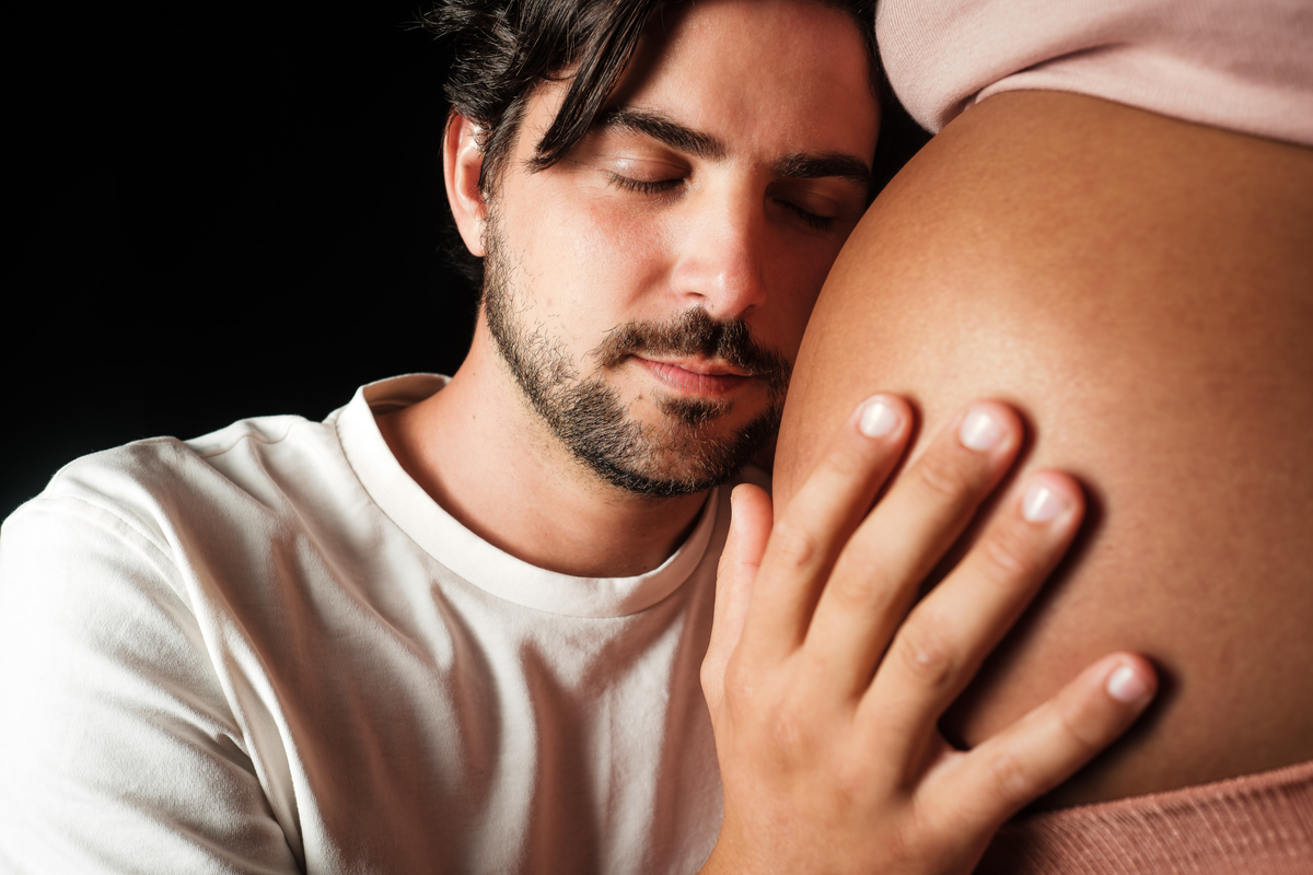 Fotografia de gestante com iluminação suave e foco na barriga. Ensaio de grávida em estúdio com roupas de tecido leve.
Pai encostado na barriga delhos fechados.