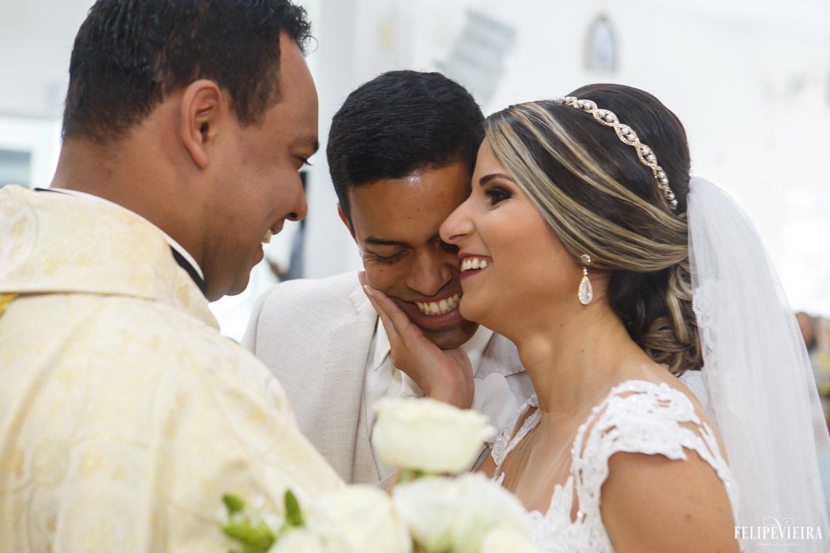 noivos sorrindo e noiva com a mão sobre o rosto de seu marido no altar em feita por Felipe Vieira fotografo de casamentos em guarapari e grande vitória