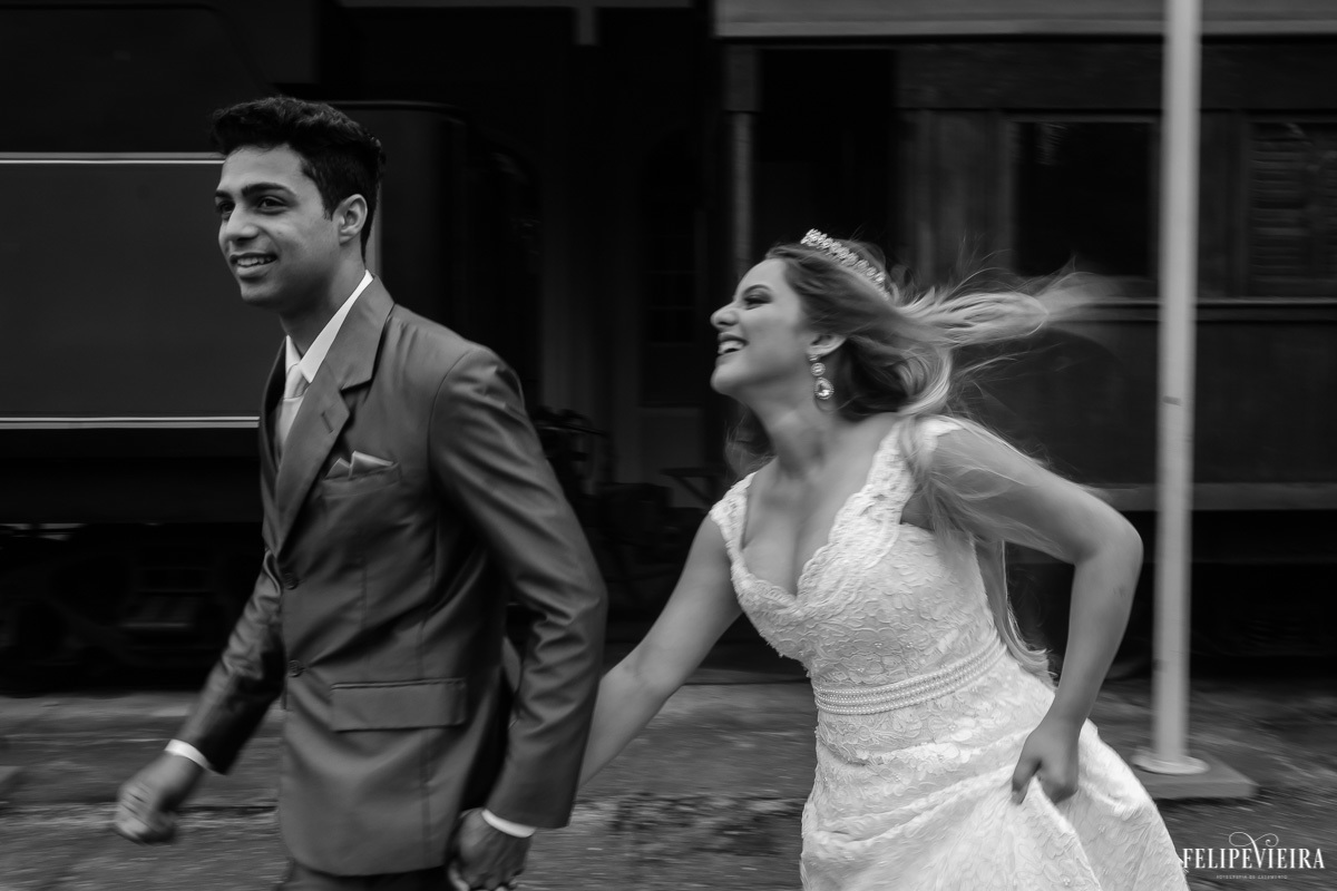 noivos correndo e sorrindo em frente a um trem e sorrindo em foto em preto e branco feita por Felipe Vieira fotografo de casamentos em guarapari e grande vitória