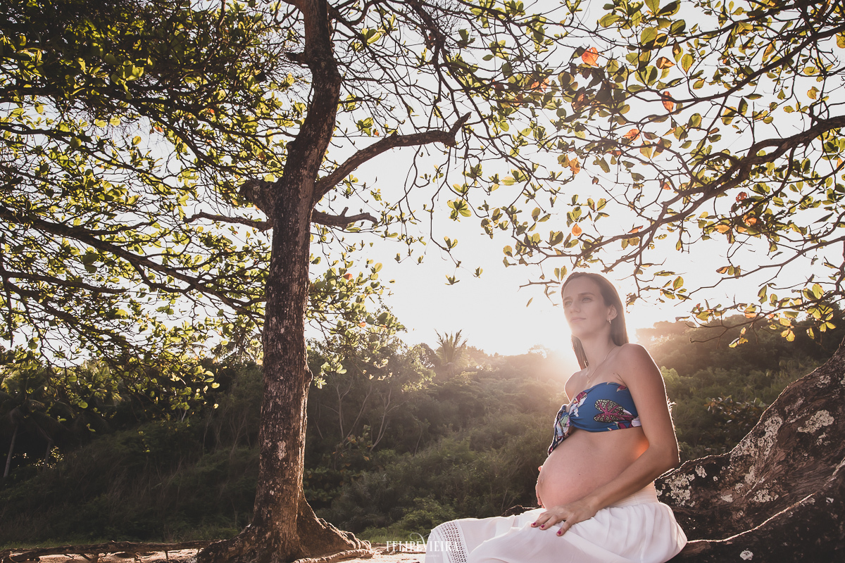 gestante durante por do sol com cropped azul e saia branca sentada na castanheira na praia em Guarapari ensaio de grávida
