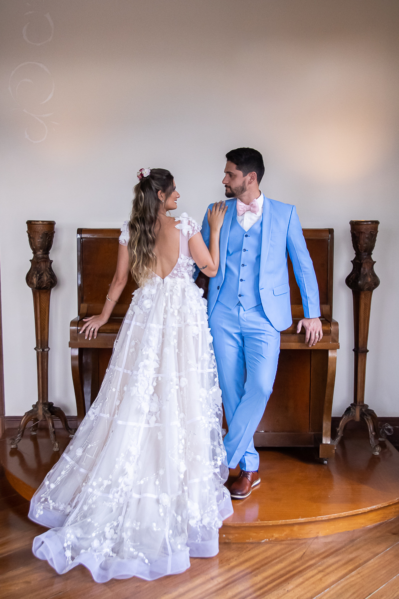 noivo com terno azul e gravata rosa apoiado no piano olhando para noiva que esta de costas com vestido estilo americano