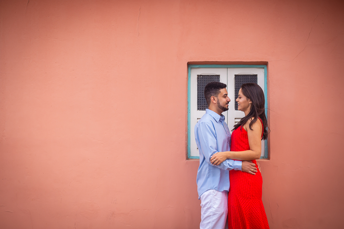 noivos em frente a uma janela com a noiva de vestido vermelho abraçando o noivo com camisa azul