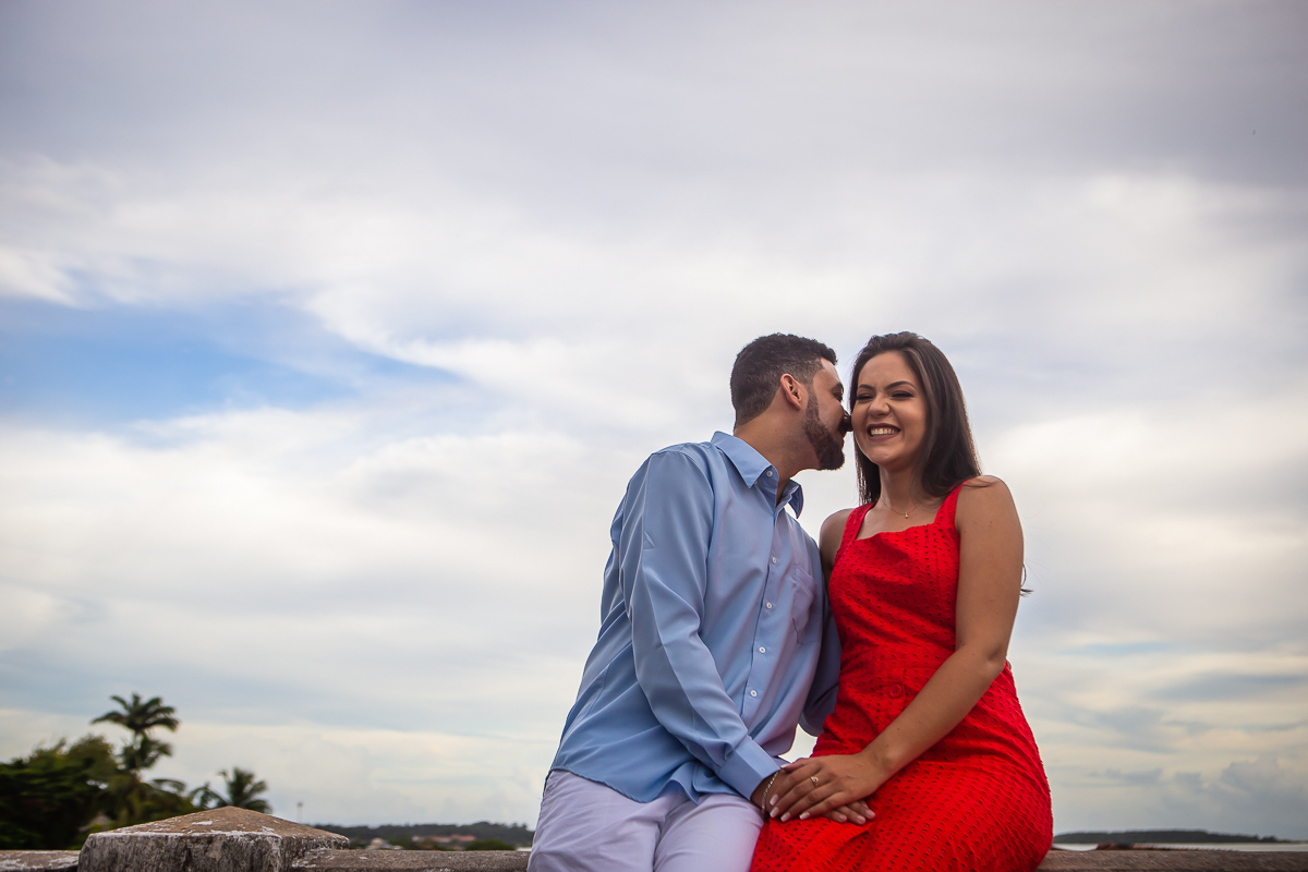 noivo falando no pé do ouvido da noiva com vestido vermelho e o céu nublado no fundo