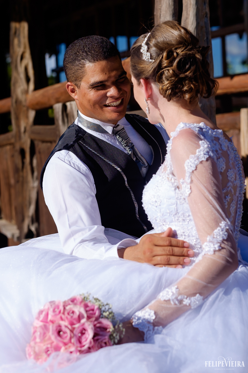 casal sentados na frente da lagoa da pousada pedra azul noiva com buquê rosa ele de frente ela de costas detalhes no fundo em madeira fotos do fotografo felipe vieira fvfotografo de guarapari