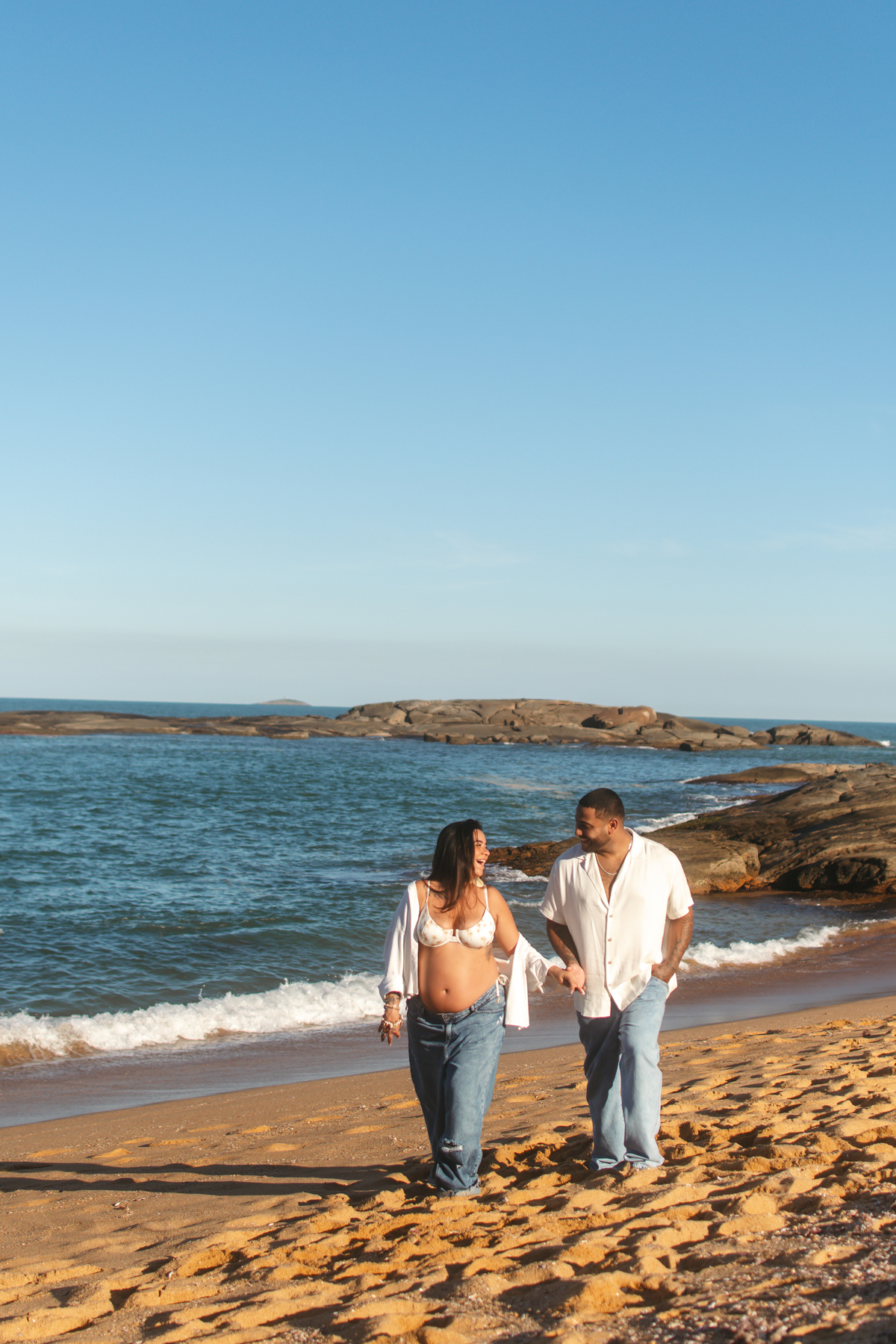 Casal caminhando pela praia durante ensaio de gestante ao entardecer.