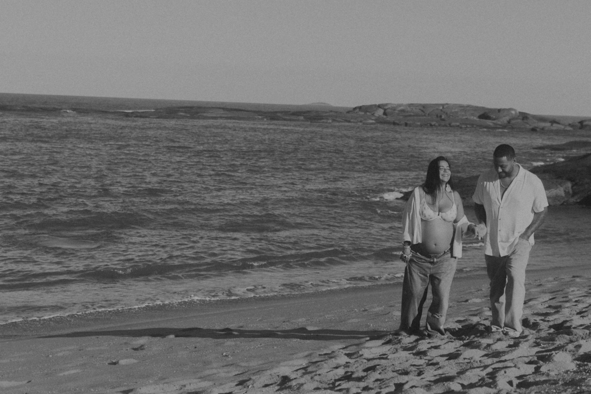 Casal caminhando pela praia durante ensaio de gestante ao entardecer em preto e branco.